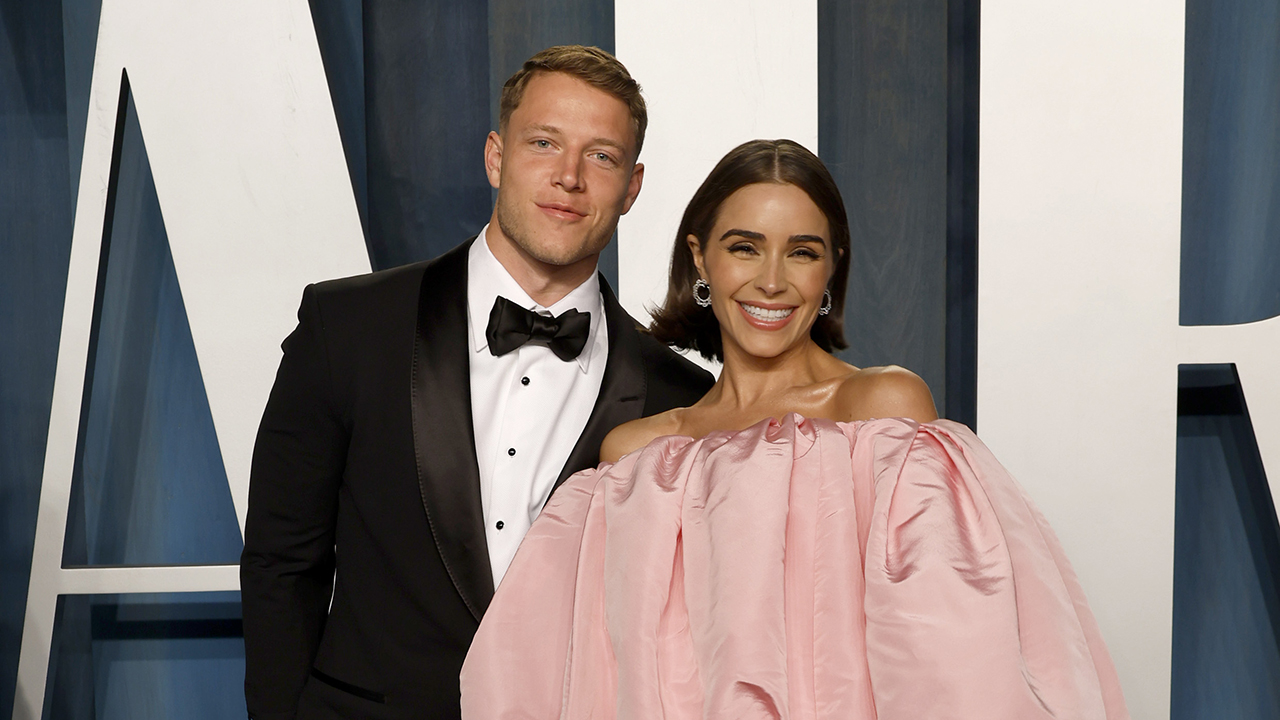 Christian McCaffrey wife. Christian McCaffrey in a tuxedo and Olivia Culpo in a pink puffy dress with a blue and white background.