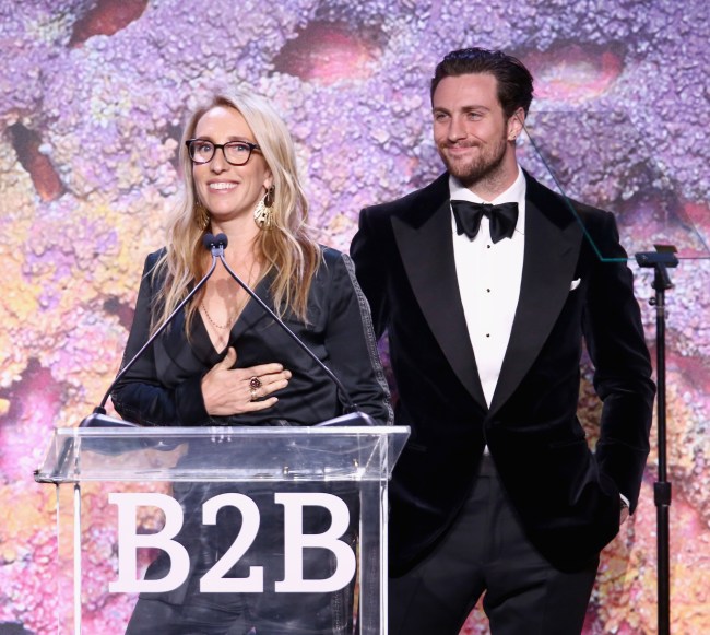 Aaron Taylor-Johnson, a white man wearing a tuxedo, standing behind Sam Taylor-Johnson, a blonde white woman wearing a black dress, who is standing in a podiun and a purple and orange background