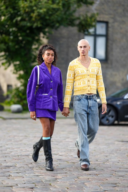 A guest (L) wears silver earrings, a neon purple velvet jacket, a matching purple velvet short skirt, a white shiny leather shoulder bag, blue high socks, black shiny leather pointed knees boots / high boots, a guest (R) wears gold necklaces, a pale yellow and white latte print pattern wool buttoned pullover, blue denim large pants, brown shiny leather ankle boots , outside Ganni, during the Copenhagen Fashion Week Spring/Summer 2024 on August 10, 2023 in Copenhagen, Denmark.