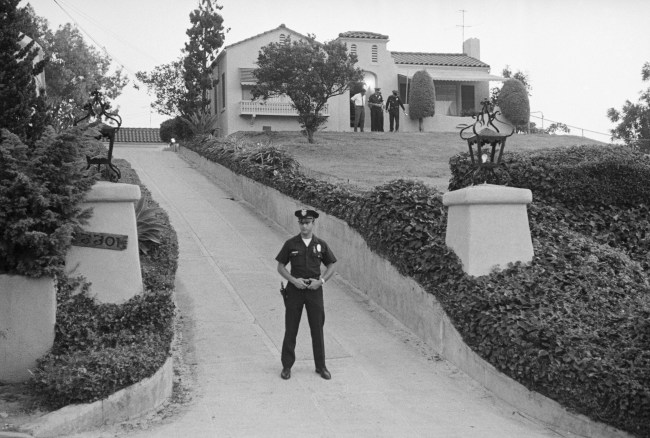 A police officer blocks the driveway while other officers search in front of the house where a middle-aged couple was stabbed to death, late, August 10. There were striking similarities between the double murder of Leon La Bianca, 44, and his wife Rosemary, 38, and the mass murders of actress Sharon Tate and four other persons the day before, and police eventually connected the killings to the "Manson Family".