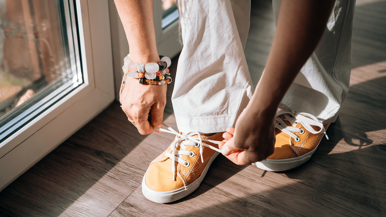 A woman in white trousers tying her shoelaces on an orange sneaker.