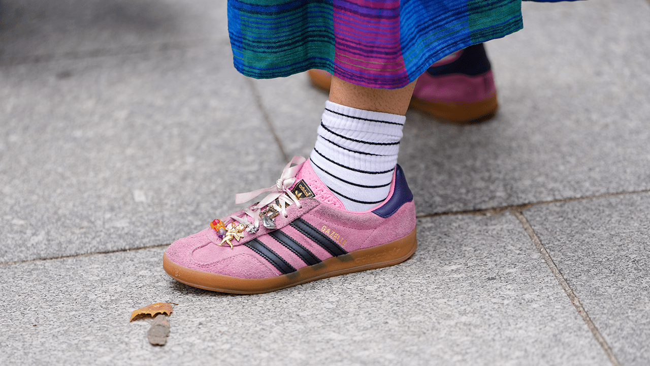 A guest wears a skirt with a vibrant blue and purple striped pattern. The footwear consists of Adidas Gazelle pink suede sneakers with black stripes and a gum sole, featuring decorative charms on the laces. White socks with black horizontal stripes are visible above the sneakers, outside Dior, during Paris Fashion Week - Menswear Spring/Summer 2026, on June 27, 2025 in Paris, France.