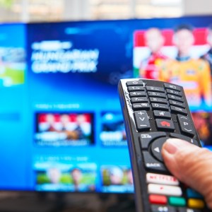 A finger pressing a button on a TV remote with a blurry TV in the background