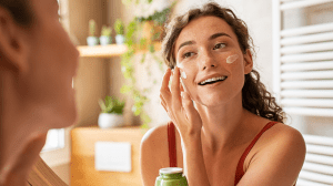 Woman caring of her beautiful skin face standing near mirror in the bathroom. Young woman applying moisturizing cream on her face during morning routine. Smiling natural girl holding little green jar of ecological cosmetic cream.